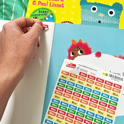 Mão aplicando ministickers em um livro infantil, ao lado de uma cartela de etiquetas