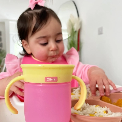Bebê com laço rosa segurando um copo de treinamento com um ministicker com o nome Olivia